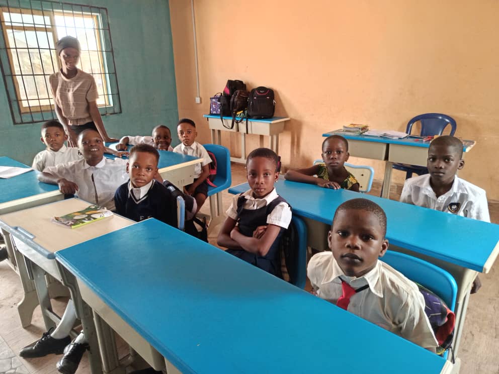 A class at long blue-topped desks, their teacher standing behind them by the window.