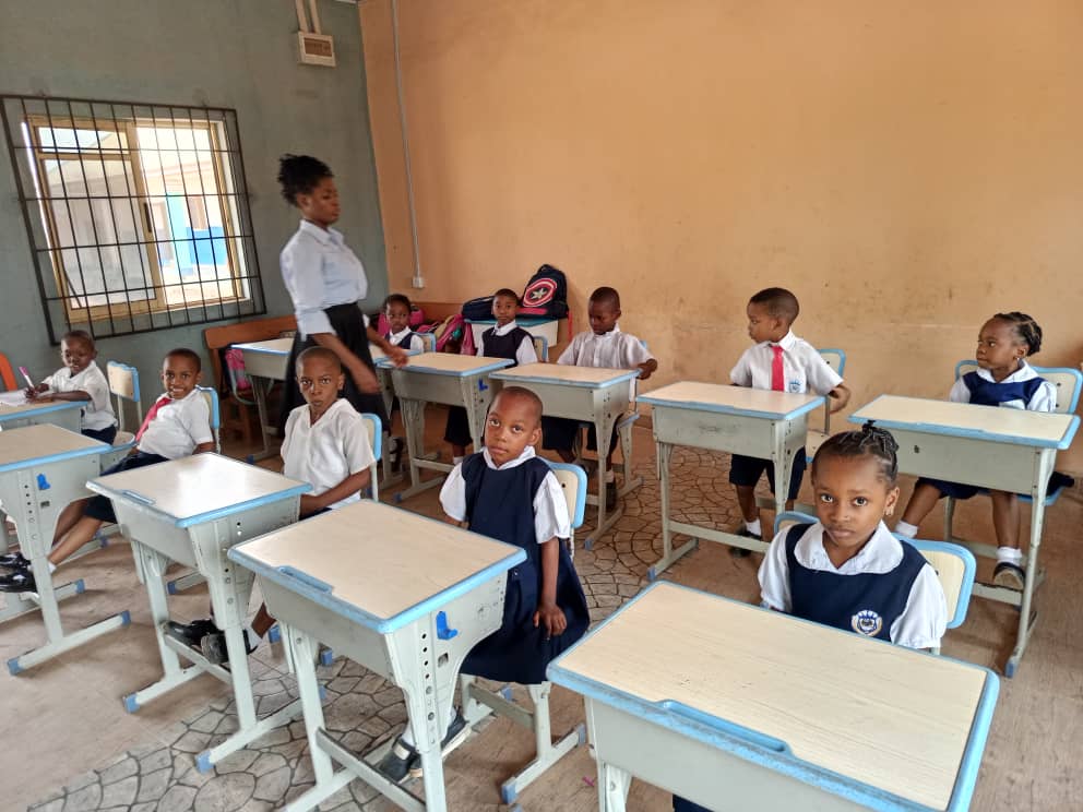 Pupils at two-seater desks turning to face the camera, with their teacher standing by the window.