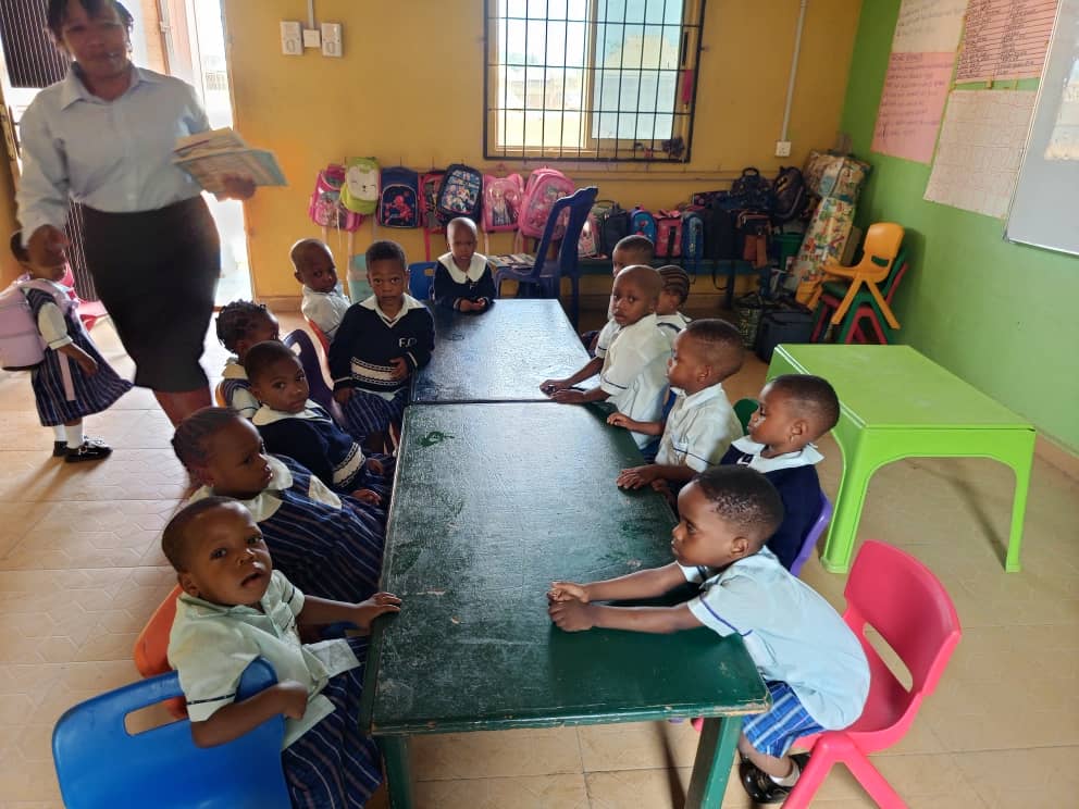 A teacher carrying an open book past a long table of very young children, with backpacks hung along the wall.