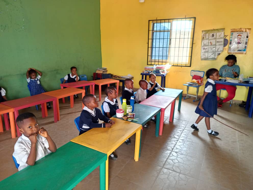 The early years room: small children at low red, green and yellow tables, with lesson charts on the wall behind them.
