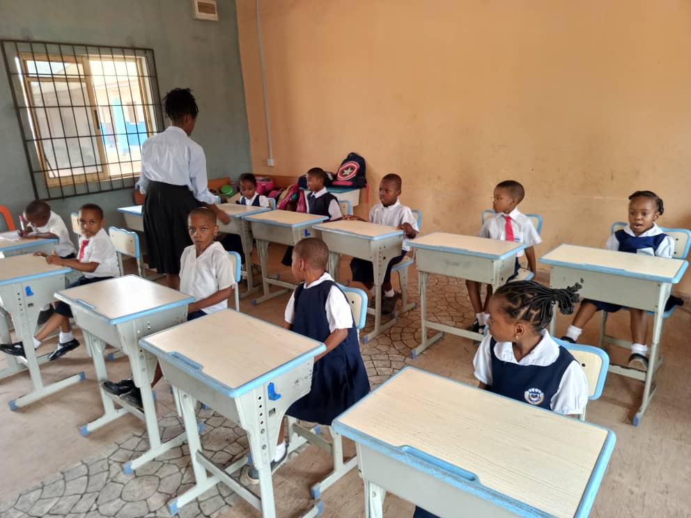 A teacher leaning over a pupil's desk at the back of a classroom while the rest of the class looks up.